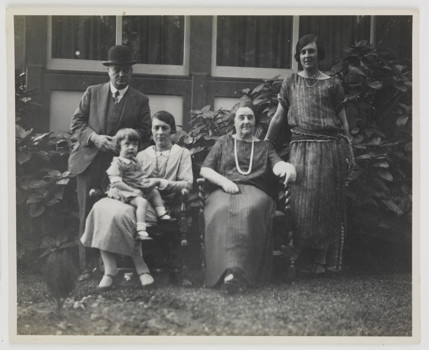 Family posing for group portrait in garden outside house. Shrub behind them. Two women are seated, one with a child on her lap. Man in hat stands at left and younger woman in dress stands at right.