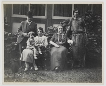 Family posing for group portrait in garden outside house. Shrub behind them. Two women are seated, one with a child on her lap. Man in hat stands at left and younger woman in dress stands at right.