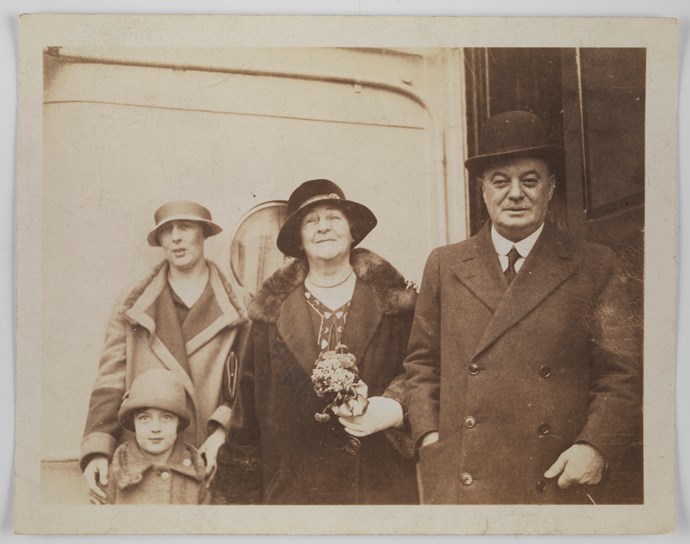 Man, two women and a young girl stand smiling on deck of ship. Woman in middle holds a posy of flowers. The women wear fur-trimmed winter coats while the man also wears a winter coat. They all wear hats.