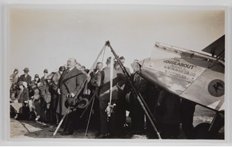 Crowd of men and women gathered at back of plane, with man speaking next to a large slingshot device. The plane has writing on the side reading: 'THE/ ROUSEABOUT / Presented to / The AUSTRALIAN AERO CLUB N.S.W / SECTION / By J.J. ROUSE ESQ.'