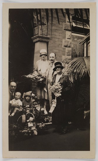 Group of people with baskets of floral arrangements around them, on the steps of a large stone building. Two women and a man stand at right, an older man and two small children sit on the stairs. The two women are holding bouquets of flowers. The women and the young girl are wearing hats. A large fern is at the right side of the group.