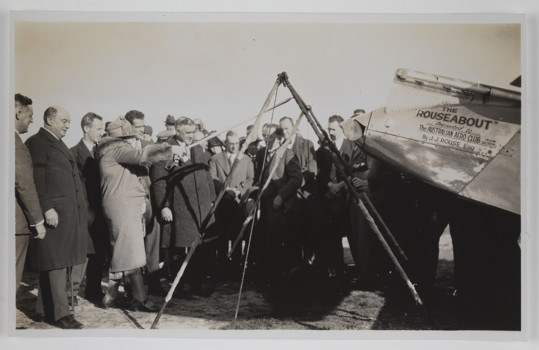 Crowd of men and women gathered at back of plane, with woman stretching out a large slingshot device. The plane has writing on the side reading: 'THE/ ROUSEABOUT / Presented to / The AUSTRALIAN AERO CLUB N.S.W / SECTION / By J.J. ROUSE ESQ.'