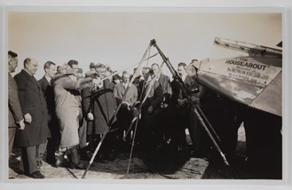 Crowd of men and women gathered at back of plane, with woman stretching out a large slingshot device. The plane has writing on the side reading: 'THE/ ROUSEABOUT / Presented to / The AUSTRALIAN AERO CLUB N.S.W / SECTION / By J.J. ROUSE ESQ.'