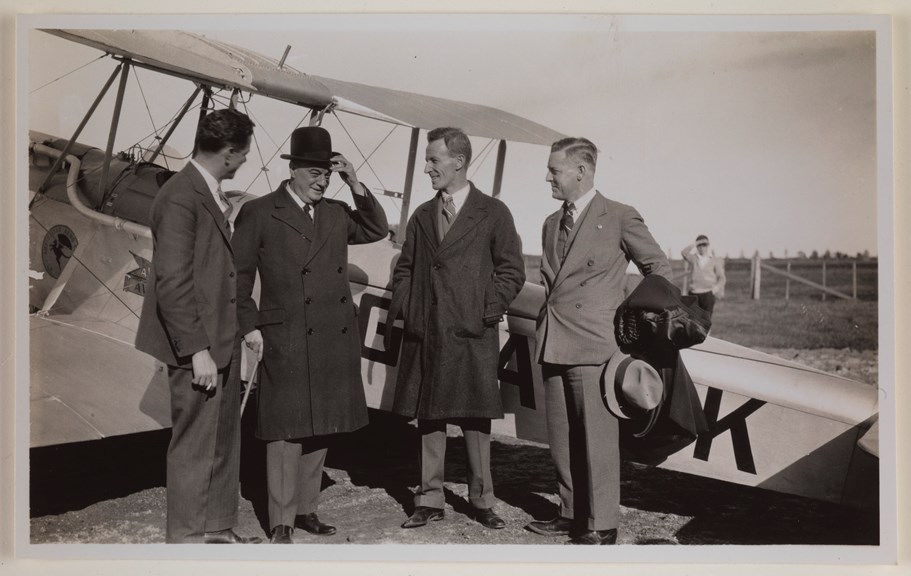 Four men smiling and talking next to a biplane. Man second from left holds a walking cane and wears a bowler hat. The two men in the middle are wearing winter coats. Man at far right holds coat and hat. Man at left has cigarette in his hand.
