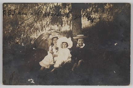 Three children wearing hats, sitting on a grassy bank underneath some trees. One girl is holding a parasol.