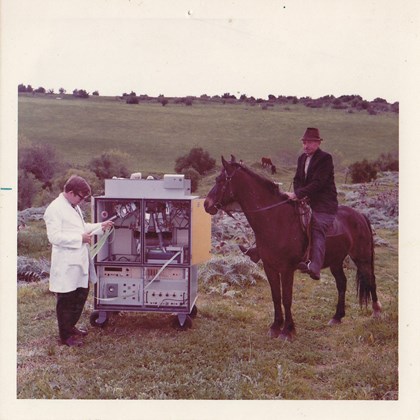 Man in labcoat with machine in field, watched by man on horseback