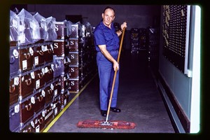 Man with a broom looks directly ahead, There storage case behind him