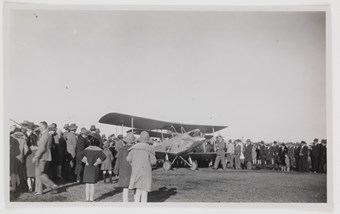 Large crowd gathered behind a small biplane parked on a grassy lawn
