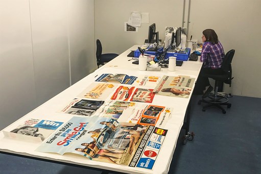Woman sitting at a computer doing data entry with a variety of colourful posters lying on the table in the foreground