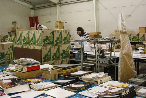 Woman standing in warehouse at back of the space, surrounded by packing boxes with books and published material on tables in the foreground