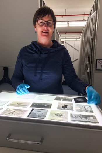 Woman in blue jumper and blue gloves standing in front of a light box with negatives on it and cabinets in the background