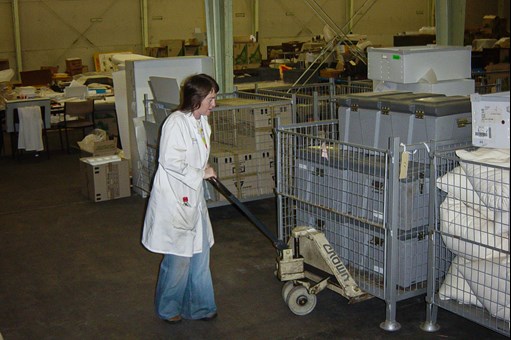 Woman in jeans and white laboratory coat pushing pallet of plastic crates in a warehouse
