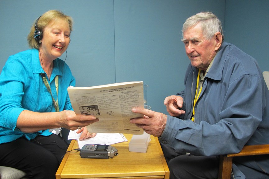 A woman with headphones on and a man in a blue jacket sitting at a table, holding a booklet in between them with a recording device on the table below