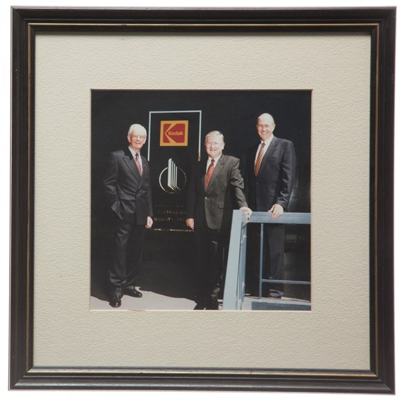 Three men in suits stand in front of a black and silver plaque, with the red and yellow Kodak 'K' symbol on top and the Quality Prize logo and text on the bottom. Photograph is mounted on white card in a wooden frame painted black and gold.