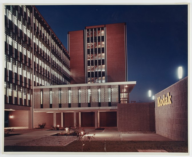Building 8 in the newly constructed Kodak Australasia Pty Ltd factory complex in Coburg, circa 1965. Exterior view of a multi-storey building at night, with two sections of the building converging in an 'L' shape. Left section of the building has fewer storeys. At ground level there is an underground car park area. To the right is a low lying wall with "Kodak" signage. In the foreground is paved and grassed areas.