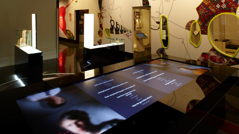 A view of a dark, empty gallery in ‘Identity: yours, mine, ours’ exhibition’. Some objects include a blurred face on digital screen, abstract faces on walls and lit up glass cabinets.  