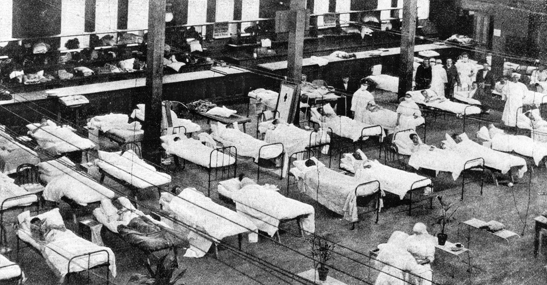 People lie in rows of sick beds in a great hall