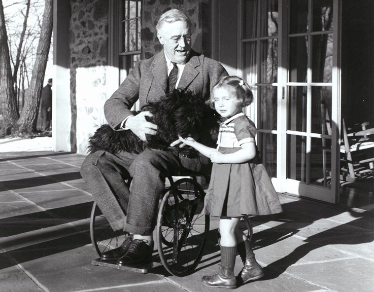 a man in a wheelchair and a dog on his lap smiles at a small girl beside him