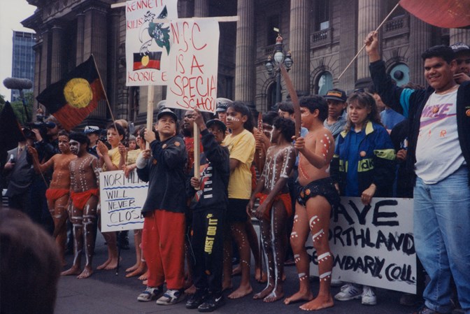 Group of people standing on the steps of parliament house holding placards and aboriginal flags
