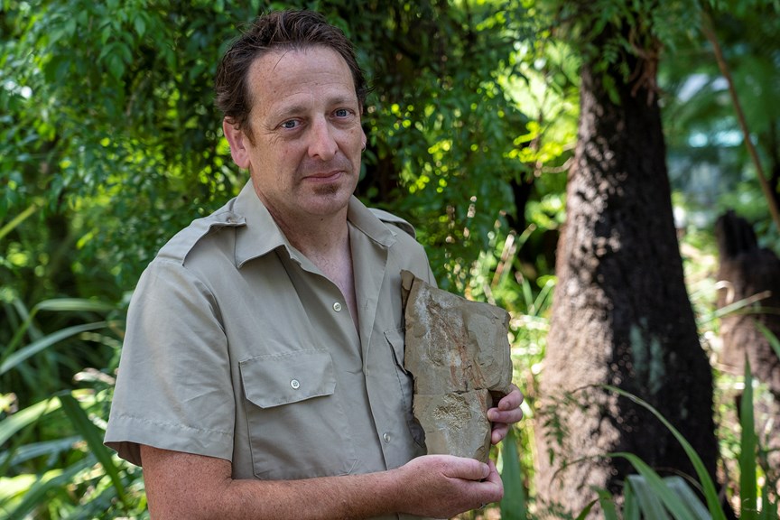 A man in a khaki shirt holds a slab of rock containing a fossil