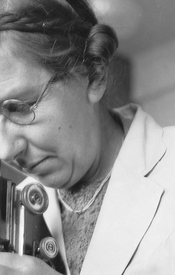 a black and white photo of a woman wearing glasses and a white coat, looking into a microscope