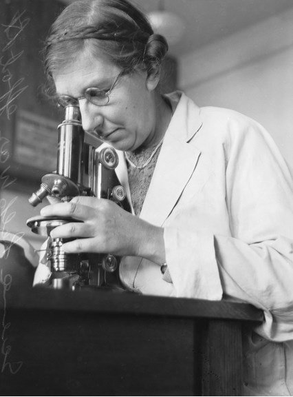 a black and white photo of a woman wearing glasses and a white coat, looking into a microscope