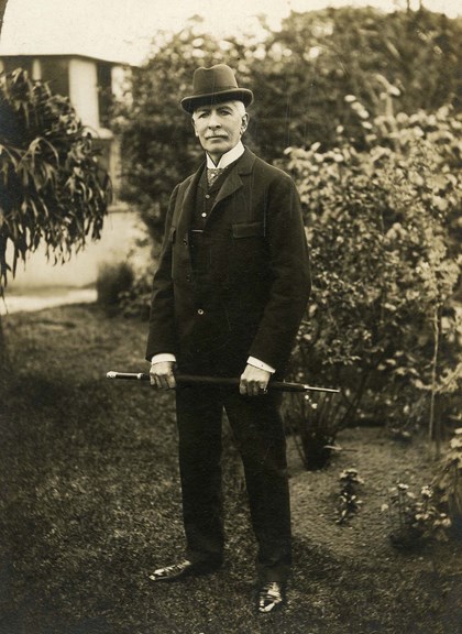 a black and white portrait photo of a man wearing a suit and hat standing in a garden, holding a walking stick