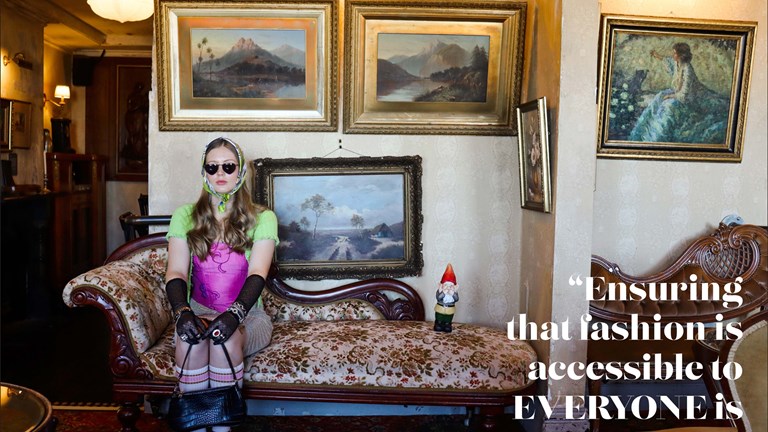 A girl sits on a chaise lounge, wearing sunglasses, a scarf, and long black gloves. Behind her hang several paintings of landscapes. A large quote overlaid on the image reads “ensuring that fashion is accessible to everyone is paramount to me”.