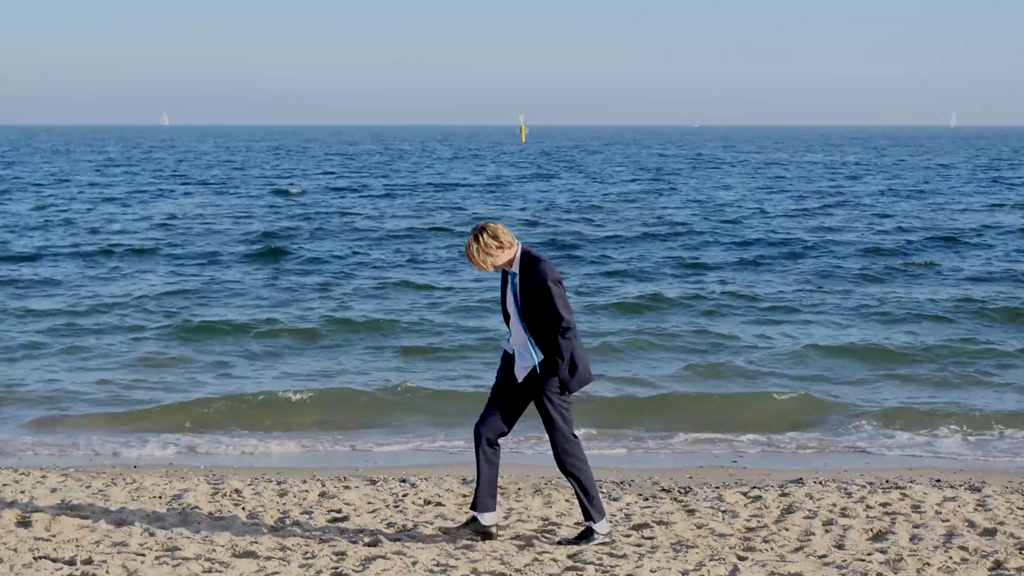 A person is walking on a beach, wearing a suit. Their hands are in their pockets and they are looking down towards the sand.