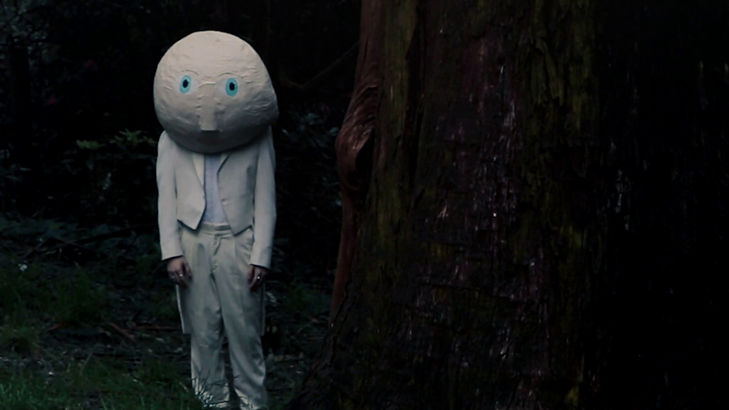 A figure stands in dark woods, wearing a white suit. They have a large spherical head made of paper mache, with two large blue eyes.