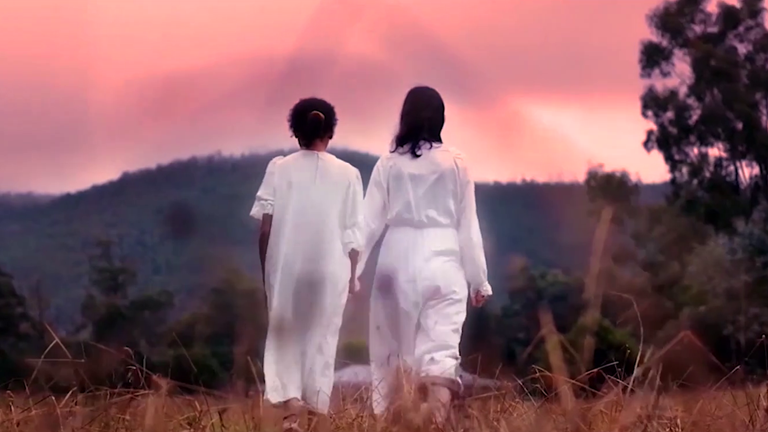 Two young women with dark hair wear long white dresses and walk closely together through an open field. They are surrounded by a natural landscape. The sky is pink and purple. 