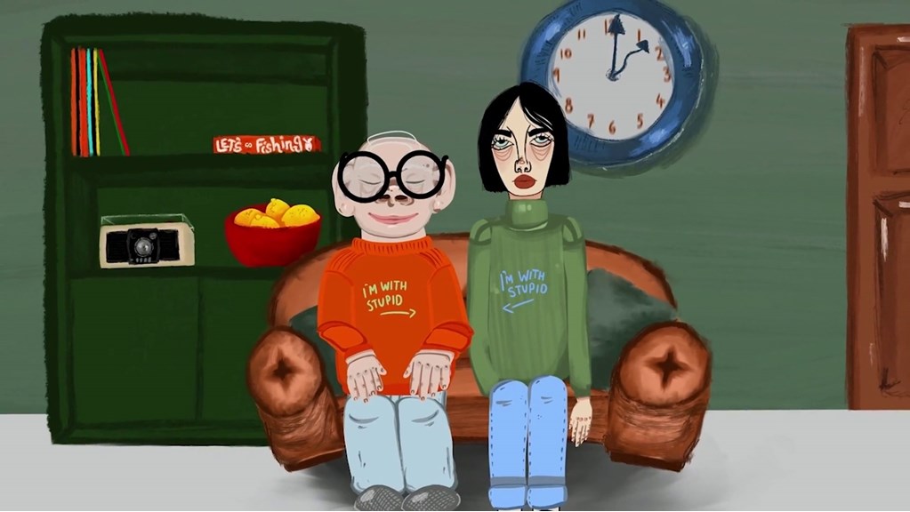 A still from an animated film, showing two people sitting together on a couch. They wear matching jumpers that read ‘I’m With Stupid’, pointing to one another. A wall clock and bookshelf are in the background.