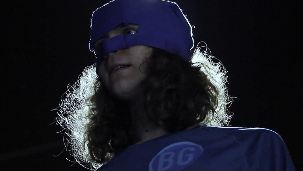 A young man with long hair wears a blue superhero mask and matching top. A cool light highlights his hair from behind and he has a surprised expression on his face. 