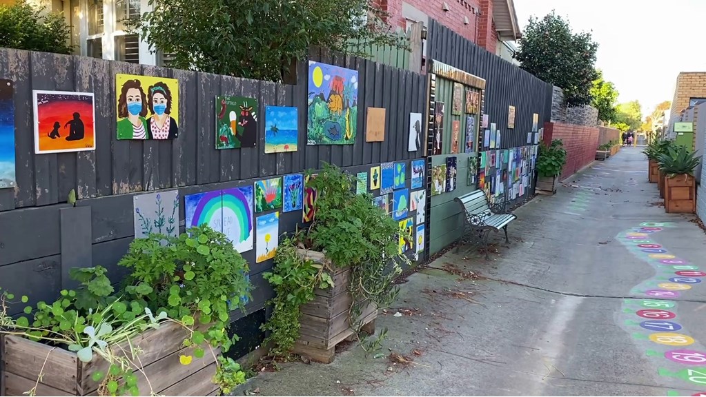 A laneway full of artworks; paintings are mounted on fences, and there are colourful numbers applied to the ground. Planter boxes full of greenery are visible along the length of the laneway.