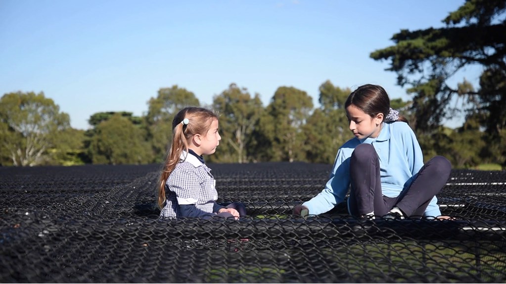 Two young girls in their school uniform sit together on top of a wire structure. The younger of the two girls faces the older and the older faces down. There are large trees in the background.