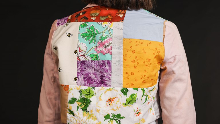 The back of a long-sleeved jacket is shown, with various colourful patterned panels, including florals, bees, and farm animals. The sleeves of the jacket are a light pink.