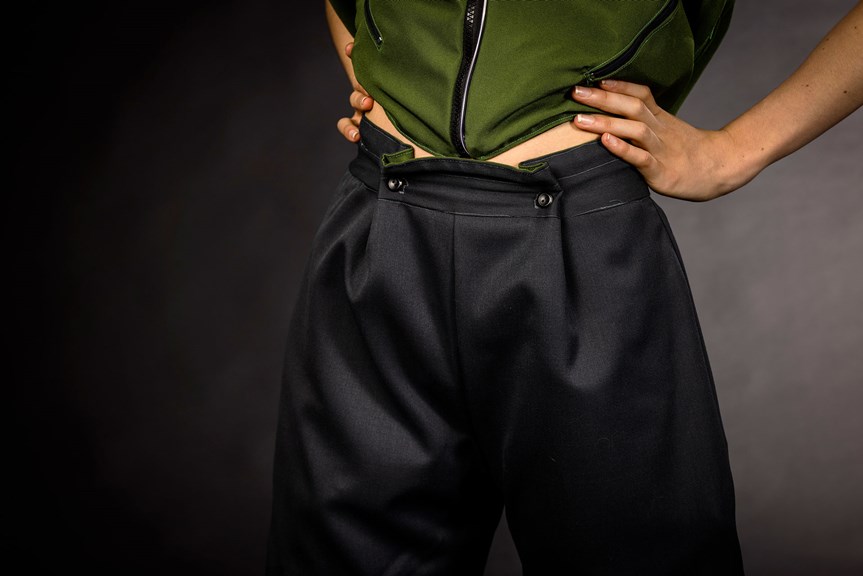A close-up photo shows a person with hands on their hips, wearing dark coloured trousers and a green top. The trousers are fastened with two buttons, and the top has zip closures. 