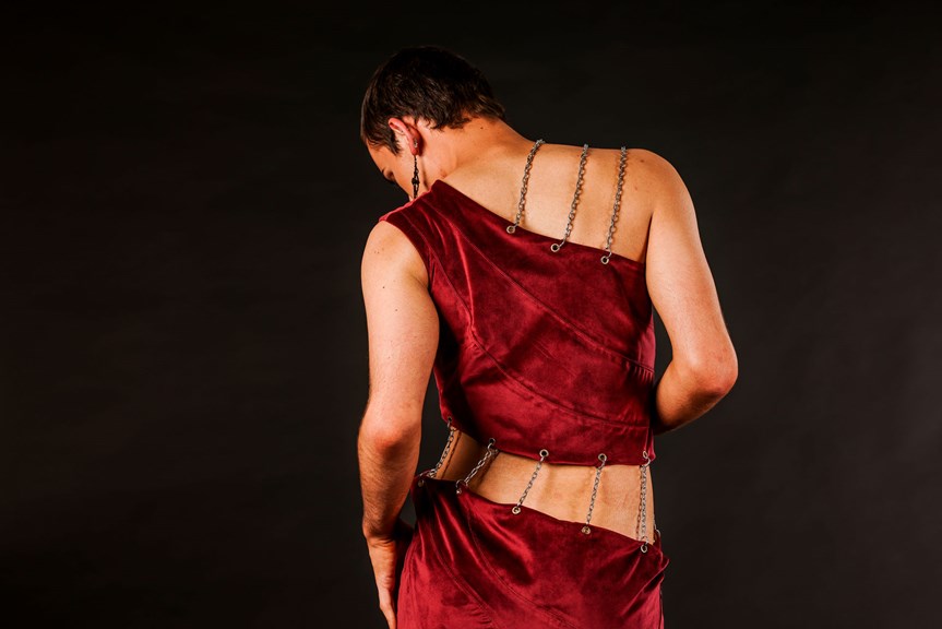 A person is shown facing away from the camera, wearing a red one shoulder garment. Two panels of the garment are visible, connected by chains. The wearer’s skin is visible in the gap between the panels.