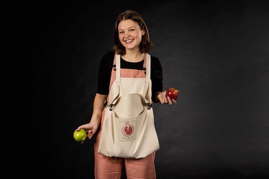 A person smiles, wearing overalls and a bag that hangs in front of them. The bag has a pomegranate logo and text reading ‘Oz Fruit Orchard’. The person holds an apple in each hand.