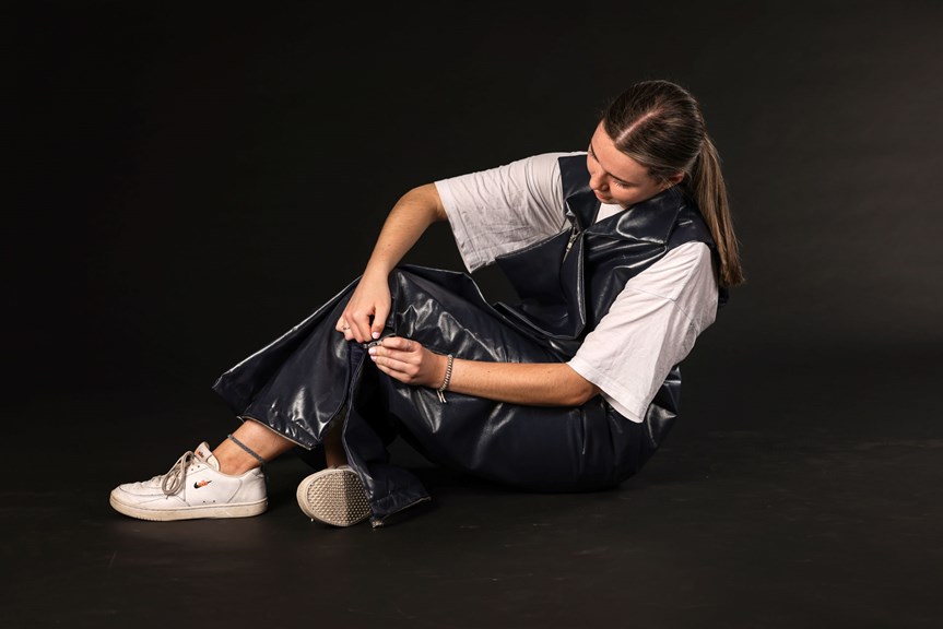 A person sits on the ground, wearing a floor length, short-sleeved jacket made of a shiny black material. They are looking down at the garment, zipping it up.