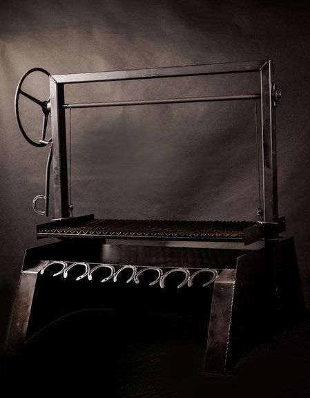 A dark-grey steel barbeque grill is pictured against a dark grey backdrop. The large barbeque has an industrial steam-punk aesthetic, with a row of horseshoes along the front and a wheel for raising and lowering the grill.