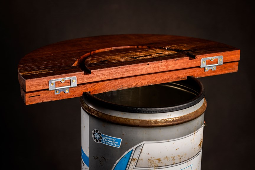 Two semi-circles of reddish wood make up the top of the table; the photograph shows they are hinged so the tabletop can be folded in half. They rest on top of a rusty salvaged metal drum with a blue and white label reading ‘Internal Corrosion Protection’.