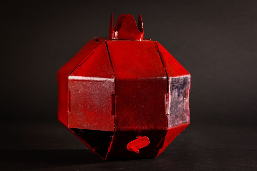 The image features the red Pomegranate against a black backdrop. The 3D octagonal shape reflects the shape and segments of a pomegranate fruit. It is made of intersecting painted acrylic sheets.