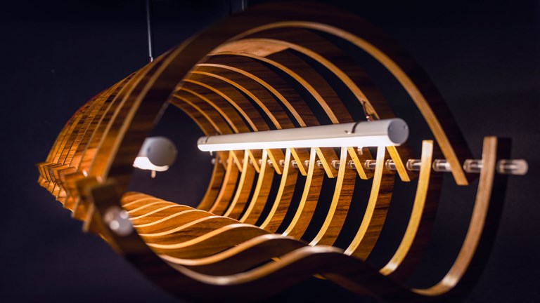 A hanging tubular lighting feature made up of many intersecting curved pieces of wood. Two LED tubes run down the centre, their light making the wood glow.