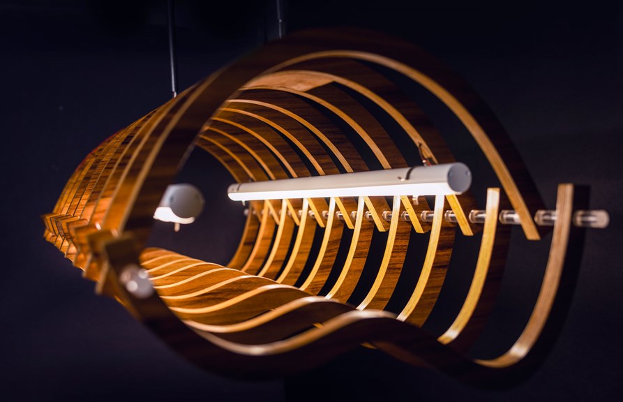 A hanging tubular lighting feature made up of many intersecting curved pieces of wood. Two LED tubes run down the centre, their light making the wood glow.