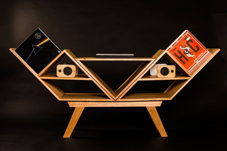 The image showcases a light-timber low shelving unit with balanced geometric shapes and angular tapered legs. A record player, speakers and music records are shown in situ on the shelves.