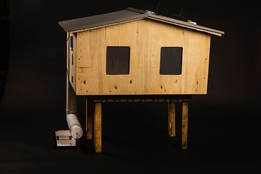 A large wooden chicken coop is suspended on four legs. It has two windows and a corrugated tin roof. A PVC pipe and wires are connected to the left side of the coop.