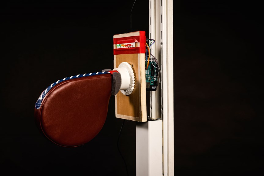 A leather paddle is connected to a wooden panel and white pole. There are coloured lights at the top of the panel, and it is fitted with an Arduino. 