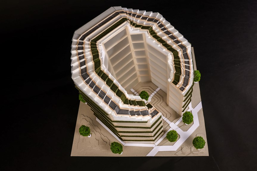 A three-dimensional architectural model of a multi-story office building. There is a void in the center, and the building is surrounded by trees. Solar panels and grass line the roof.