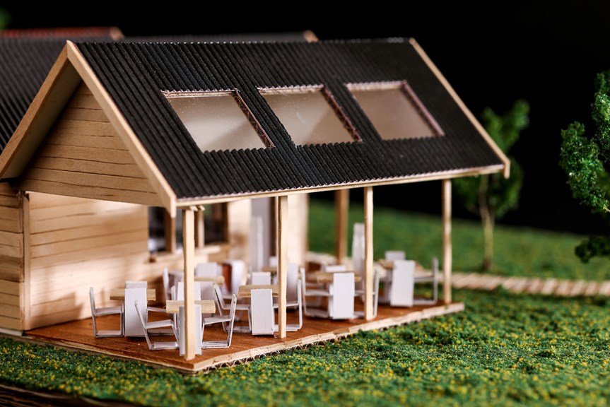 A three-dimensional architectural model of a retail store. The building has an open patio set with several tables and chairs. It sits atop grass. 